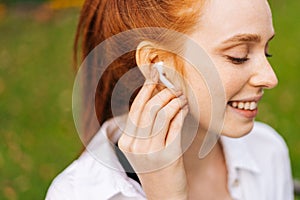 Joyful redhead female with closed eyes listening music using wireless white headphones outdoors.