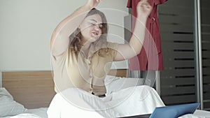 Joyful relaxed woman sitting on bed with laptop and dancing. Portrait of positive young Caucasian lady enjoying weekend