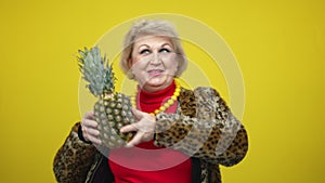 Joyful senior Caucasian woman dancing with pineapple on yellow background. Portrait of happy relaxed carefree lady