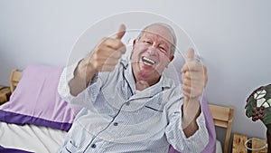 Joyful senior man, thumbs up in success, sitting on bed in pyjamas, a gesture of winner positivity in bedroom