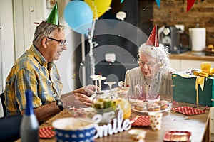 Joyful senior woman blowing candles on birthday cake, sitting at the kitchen table with her husband, celebrating her birthday