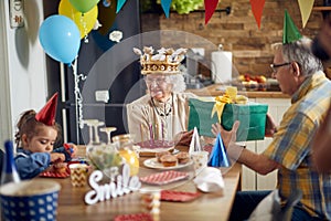 Joyful senior woman recieving a birthday gift from husband, sitting at kitchen table with their granddaughter, baloons and
