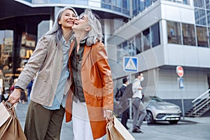 Joyful senior woman with shopping bags hug Asian lady on modern city street