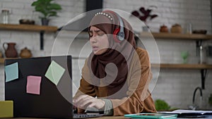 Joyful slim beautiful woman in hijab typing on laptop keyboard singing to music in headphones. Positive cheerful happy