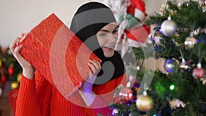 Joyful smiling beautiful woman in hijab shaking Christmas present at New Year tree in living room at home. Portrait of