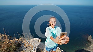 Joyful smiling girl having video chat on high cliff over sea.
