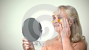 Joyful smiling lady applying eye patches looking in mirror, anti-aging therapy