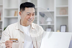 Joyful successful asian businessman working from bright home office, man using bank credit card and laptop for online