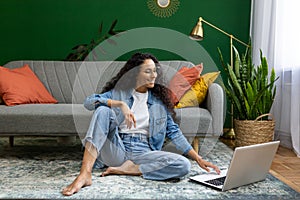 Joyful successful woman working on laptop remotely, Hispanic woman sitting on carpet on floor in living room, happily