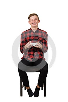 Joyful teen boy seated on chair playing video games isolated on white background. Excited adolescent holding a joystick console