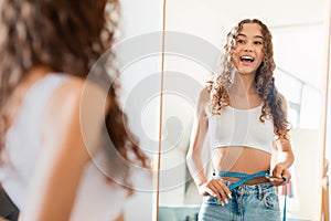 Joyful teen girl measuring her waist with tape standing indoors