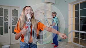 Joyful teen girl with paint roller singing dancing as blurred man painting wall at background. Portrait of happy