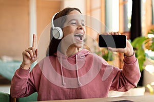 Joyful teen girl singing song and using smartphone as microphone, wearing wireless headset, having fun at home