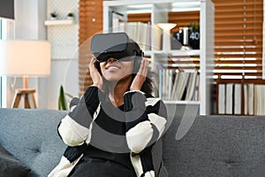 Joyful teenage asian woman experiencing opportunities of gaming in virtual reality headset while sitting ouch in living