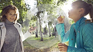 Joyful teenage caucasian girl blowing soap bubbles to her positive grandmother in the sunny autumn park. Family having