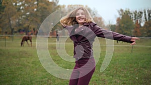Joyful teenage girl spinning smiling on autumn meadow with horse barn at background. Portrait of excited carefree