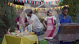 Joyful teenage girl walking in slow motion to busy group of multiethnic friends cooking barbecue and setting picnic