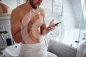 Joyful topless man texting while brushing teeth