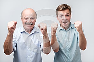 joyful two young men clench fists and shout with happiness, dressed casually, isolated on white background. Happy son and