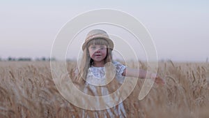 Joyful village childhood, little cute kid girl in white dress and straw hat spins touching reaped grain oat spikes in