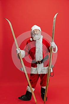 Full-length portrait of senior man wearing Santa Claus costume, holding skis isolated over red background. Winter