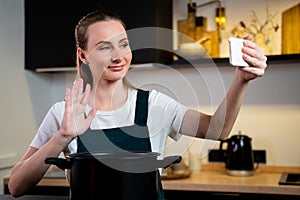 Joyful woman in an apron cooks at home and tastes a dish, shooting a video tutorial on the phone in the kitchen interior