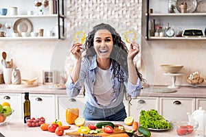 Joyful woman having fun with glasses of sweet pepper, playing with food and fooling around, cooking salad in kitchen