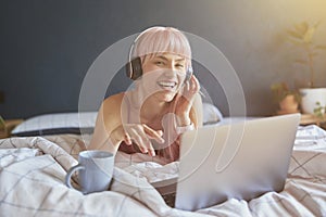 Joyful woman with headphones listens to music on laptop on bed