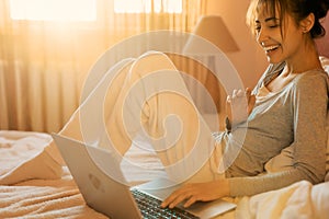 Joyful woman laughting and talking on video call on laptop computer in bed at home, sunlight in bedroom.