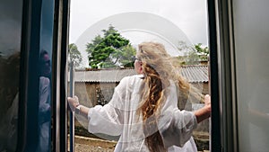 Joyful woman leans out train door, long hair flying, white shirt billowing. Carefree traveler rides through rich tropics