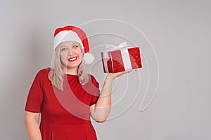 A joyful woman in a Santa hat holds a red gift box in the palm of her hand