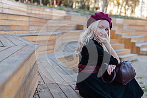 A joyful woman sits on a summer theater bench, made of wood in a burgundy coat and biret, with beautiful eyes, in the