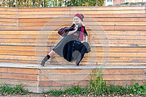 A joyful woman sits on a summer theater bench, made of wood in a burgundy coat and biret, with a beautiful smile, in the