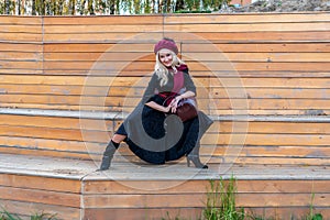 A joyful woman sits on a summer theater bench, made of wood in a burgundy coat and biret, in the fall against the blue