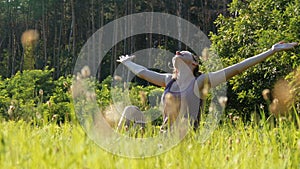 Joyful woman sitting on the green lawn happily lifts her hands up to the sky on scenic field at sunset background