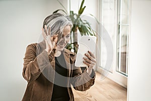 Joyful woman waves her hand using tablet computer for communication with loved ones in internet. Happy aged woman have