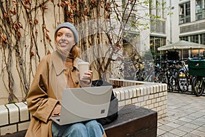 Joyful woman working on laptop and drinking takeaway coffee while sitting outdoors