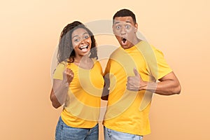 Joyful young african american couple celebrating success with thumb up and clenched fists, yellow background