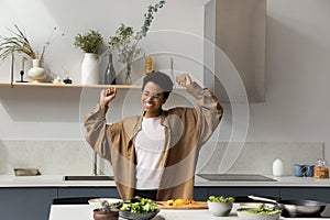 Joyful young African American woman dancing, cooking in kitchen.