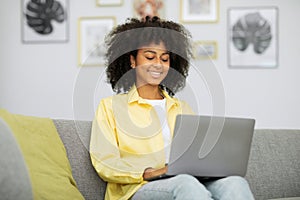Joyful young african american woman working on laptop, sitting on sofa in home office, copy space.
