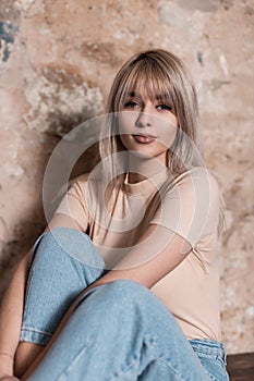 Joyful young amazing woman in a fashionable shirt in vintage blue jeans sit near an old brick wall indoors. Pretty happy girl