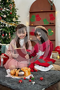 A joyful young Asian girl is reading a Christmas fairy tale story in a book to her younger sister