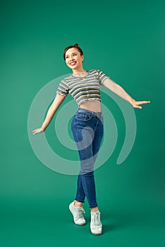 Joyful young Asian woman standing on one leg. Full length view of blissful girl dancing on green background