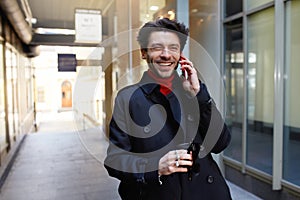 Joyful young attractive brown haired male with trendy haircut laughing happily while having phone conversation with friend,