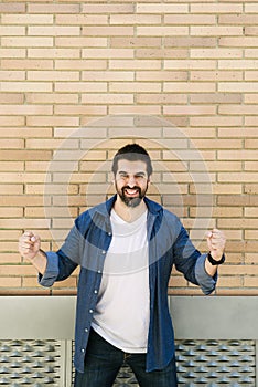 Joyful young bearded man in casual blue shirt posing on bricked wall background