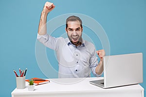 Joyful young bearded man in light shirt sit, work at desk with pc laptop isolated on pastel blue background. Achievement