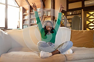 Joyful young black woman in wireless headphones listening to music, raising hands up, dancing on couch at home