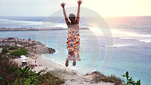 Joyful young brunette woman in long dress jumping on the rock by the ocean during sunset. slow motion. 3840x2160