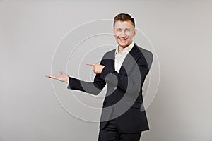 Joyful young business man in classic black suit white shirt pointing index finger hand aside isolated on grey wall