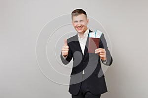 Joyful young business man in classic suit showing thumb up, holding passport, boarding pass ticket in hand isolated on